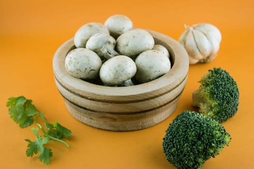 button mushrooms in wooden bowl with garlic, parsley and broccoli on orange background.