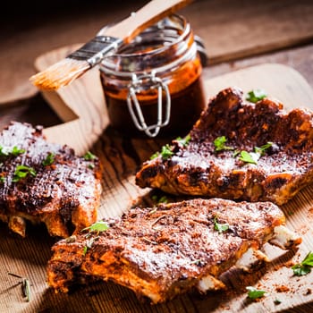 Three portions of barbecued ribs in a spicy marinade seasoned with pepper and fresh herbs being prepared in a country kitchen with a jar of basting sauce