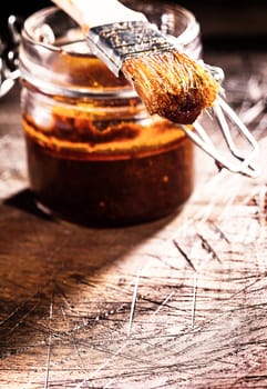 Glass jar of homemade spicy basting sauce and a brush on an old scored wooden chopping board in a rustic kitchen