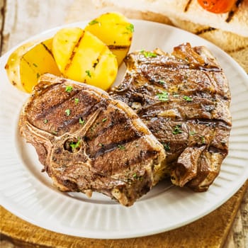 Close up of a serving of two succulent grilled barbecued lamb chops seasoned with fresh herbs served with potatoes at a summer picnic