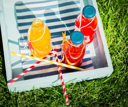 Bottles of fresh pure fruit juice with colorful striped straws matching the color of the orange and berry juice served on a wooden tray outdoors on the grass at a summer picnic