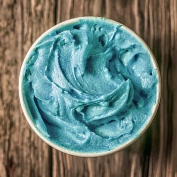 Cool refreshing turquoise blue ice cream in a takeaway tub standing on an old textured wooden table, overhead view in square format