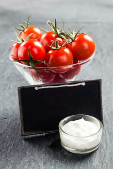 Glass bowl of fresh ripe red cherry tomatoes and savory creamy vinaigrette sauce on a slate background with a blank vintage school slate with copyspace for your text