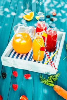 Bottles of freshly squeezed homemade citrus and berry juice with a jug of iced carrot and orange juice blend served on a wooden tray on a colorful turquoise picnic table outdoors