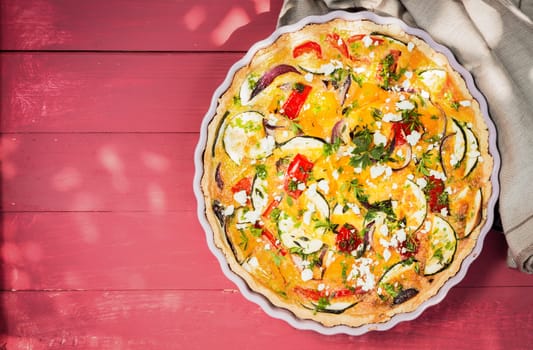 Freshly baked savory quiche with eggplant, pepper, tomato and cheese seasoned with fresh herbs in a fluted pie dish, overhead view with copyspace on red wooden boards