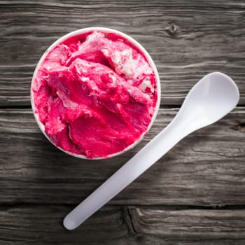 Delicious cold refreshing berry ice cream in a takeaway plastic tub with a teaspoon on an old textured wooden surface, overhead view in square format