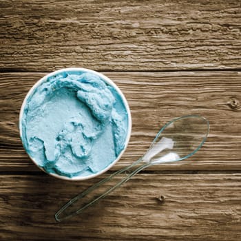 Close up overhead view of a tub of gourmet blue Italian ice cream with a plastic spoon served on an old wooden table, in square format