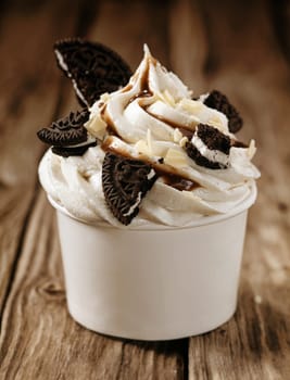 Takeaway tub filled with a swirl of vanilla ice cream with pieces of oreo biscuits and chocolate sauce, close up view on old wooden boards