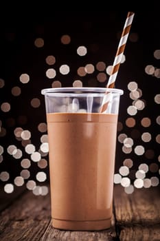 Plastic glass of delicious creamy chocolate smoothie or milkshake against a background of sparkling festive party lights