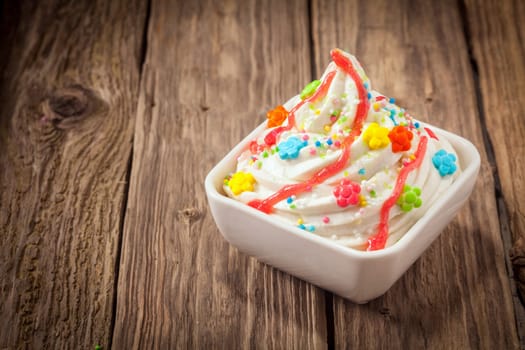 Colorful party ice cream dessert frozen yogurt with a swirl of vanilla ice cram topped with bright multicolored candy and sprinkles drizzled with syrup on an old textured wooden table with copyspace