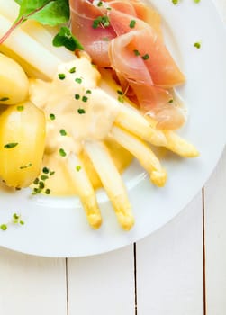 Delicious cold dinner of asparagus and prosciutto ham served with mayonnaise and boiled baby potatoes garnished with fresh chopped herbs, close up overhead view on a white plate and table