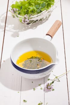 Ceramic dish of golden olive oil and chopped fresh herb salad dressing on a white wooden kitchen counter with a bowl of coriander