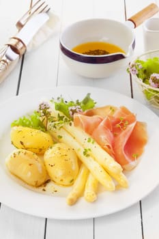 Cooked asparagus spears with Italian prosciutto ham and boiled baby potatoes served with an olive oil and herb dressing on a white wooden counter in a country kitchen