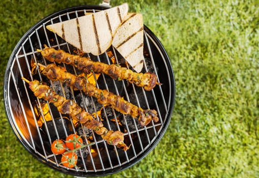 View from above of two lamb, beef or pork kebabs with slices of toast and tomatoes grilling on a BBQ fire on a portable metal barbecue during a summer picnic on green grass with copyspace
