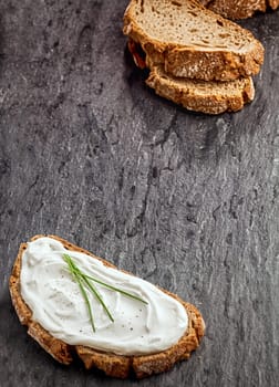 Savory cream cheese on a slice of rye bread garnished with fresh chives with additional slices of bread forming a diagonal corner border on a textured dark background with copyspace
