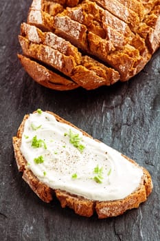 Fresh cream cheese with herbs served on a slice of wholesome rye bread viewed from above on a dark textured surface alongside the slice rye loaf