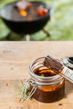 Jar of basting sauce and a basting brush standing on a wooden picnic table outdoors for the meat grilling on the BBQ in the background on a green lawn