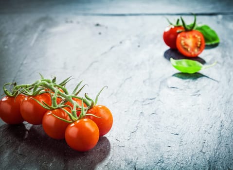 Fresh grape tomatoes on the vine in the lower corner of a dark textured wooden surface with highlight and vignetting and a halved tomato in the top corner with copyspace between