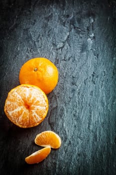 Peeled fresh clementine or tangerine with segments and a whole fruit arranged in a corner on a dark slate background with texture, copyspace and vignetting