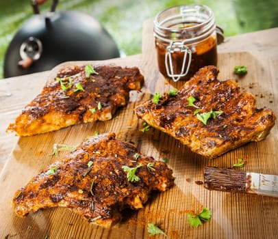 Seasoned ribs on a rustic wooden picnic table at a BBQ in the garden with fresh herbs and a glass jar of spicy marinade or basting sauce