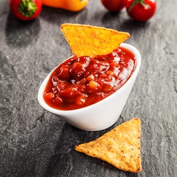 Bowl of delicious hot spicy tomato and chili salsa sauce served with triangular corn tortillas on a slate surface as a snack and appetizer