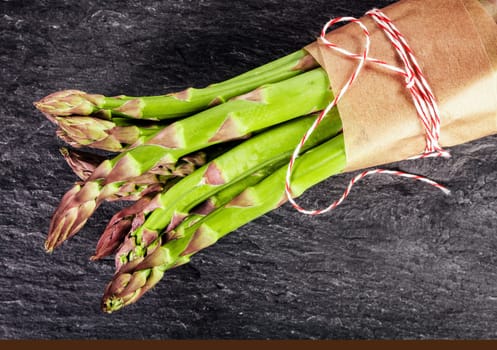 Fresh green asparagus shoots tied in a bundle with brown paper and string, close up overhead view on dark slate with copyspace