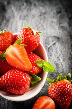 Bowl of ripe red strawberries with green stalks and one halved to show the juicy pulp, high angle view on a textured grey slate surface with copyspace