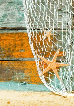 Rustic marine background with starfish suspended in a fishing net over weathered blue and orange painted wooden boards with copyspace and beach sand below