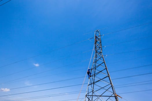 Electricians climb steel electrical tower for maintenance repairs on high powered electrical cables in blue sky.