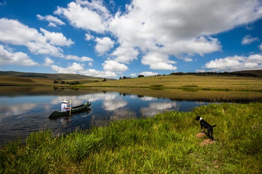 Woman paddling canoe on small mountain lake dam with glass mirror reflections of clouds and blue sky and local dogs attention on the scenic beauty landscape