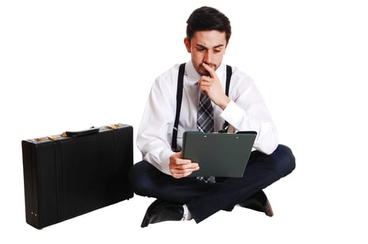 A young business man sitting on the floor looking on his notes with his.briefcase beside him, isolated on white background..