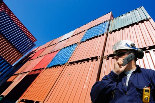 port and dock worker talking in phone with stacks of cargo containers in the background
