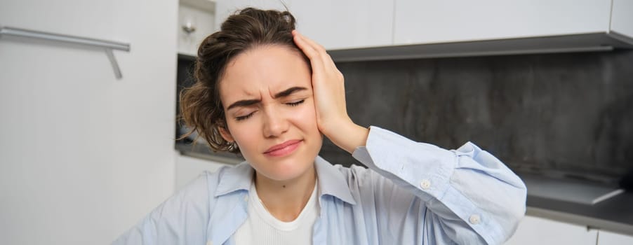 Close up portrait of brunette woman, touches her head, feels dizzy, has migraine or headache, grimaces from pain.