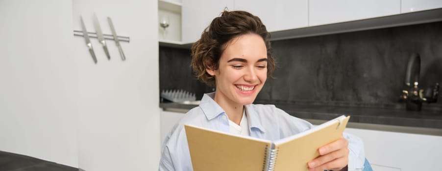 Portrait of happy brunette girl, looking at her notes, reading smth funny and laughing, sitting at home, enjoying cozy weekend.