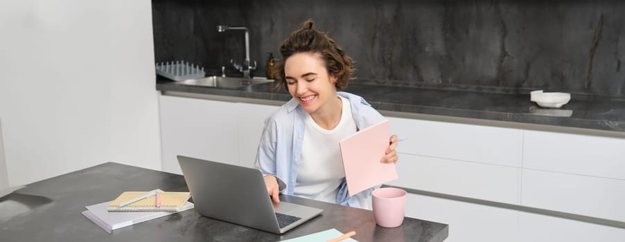 Smiling young woman laughing, video chats on laptop, connects to online work meeting, studies from home, holds her notebook, sits in kitchen.