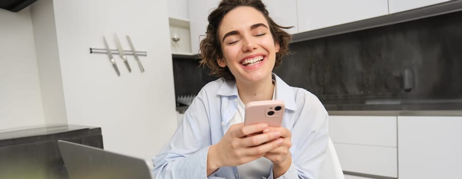 Image of young woman using her smartphone at home. Girl sits with mobile phone in kitchen and smiles.