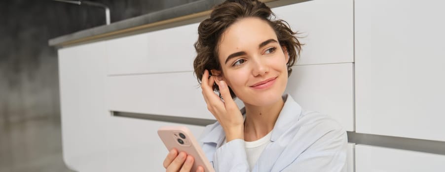 Indoor shot of happy girl with smartphone, sits on floor at home and uses mobile phone, watches video in app, chats with friends in applicatiion.