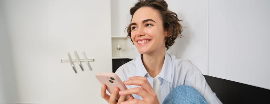 Portrait of smiling beautiful woman, sitting in kitchen with smartphone, looking happy at camera, spending time at home, using mobile phone app.