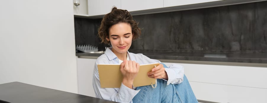 Young woman sitting in kitchen, reading her notes in notebook. Smiling girl at home, doing homework, studying.