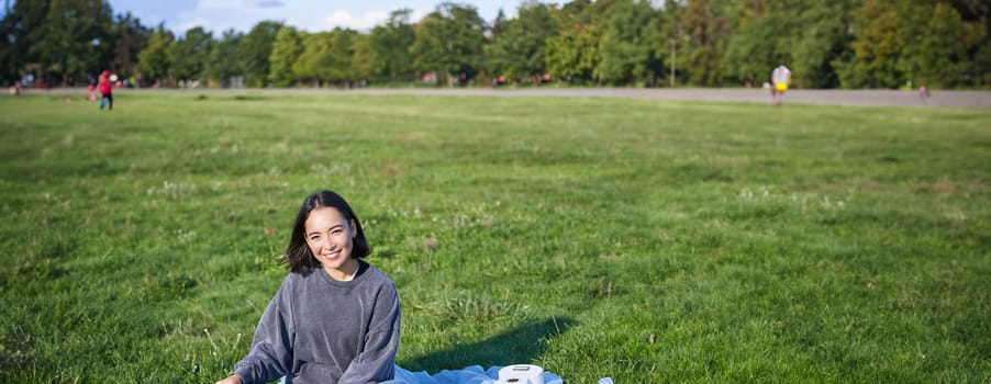 Vertical shot of smiling asian girl sitting on blanket with ukulele guitar, relaxing on sunny day outdoors, resting in park.