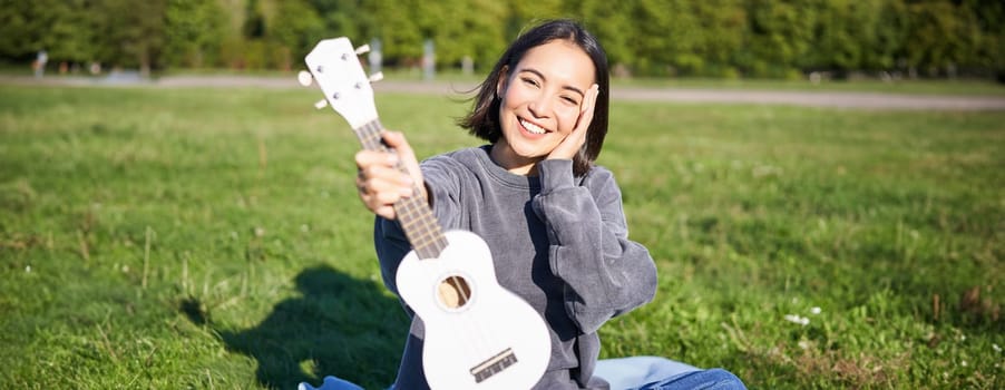 Beautiful asian girl with happy smile, shows her ukulele, sits outside in park on grass, relaxes with music.