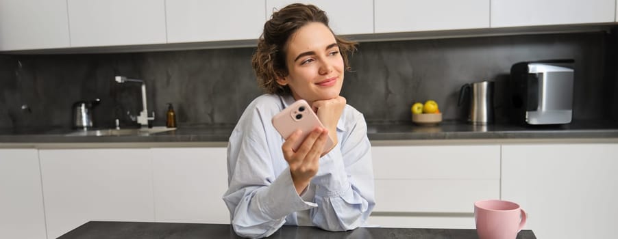 Portrait of beautiful smiling woman sitting at home with smartphone, using mobile phone app, orders groceries delivery.