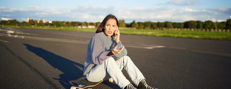 Portrait of asian girl sits on her longboard, skating on skateboard and talking on mobile phone, having conflict during telephone conversation, arguing with concerned face.
