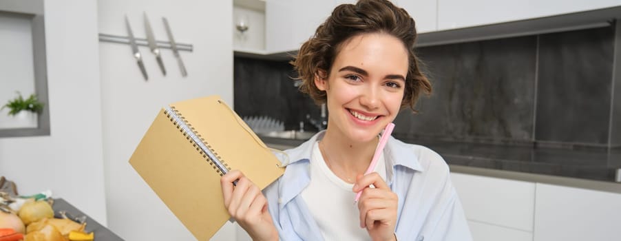 Smiling brunette womnan, holding her diary, writing down in planner, working from home on laptop, studying online and taking notes in notebook.