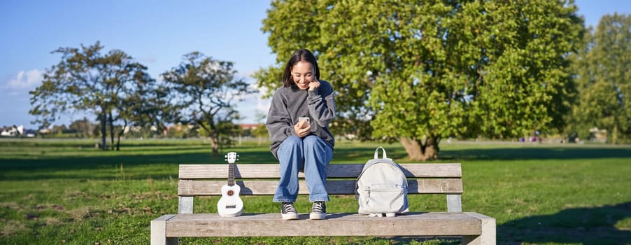 Excited girl looks at smartphone and celebrates, wins on mobile phone, sits with ukulele and backpack in park on bench on sunny day.