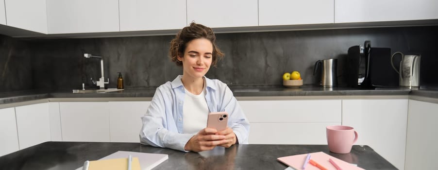 Technology and lifestyle. Young woman sits at home, uses smartphone in her kitchen and smiles.