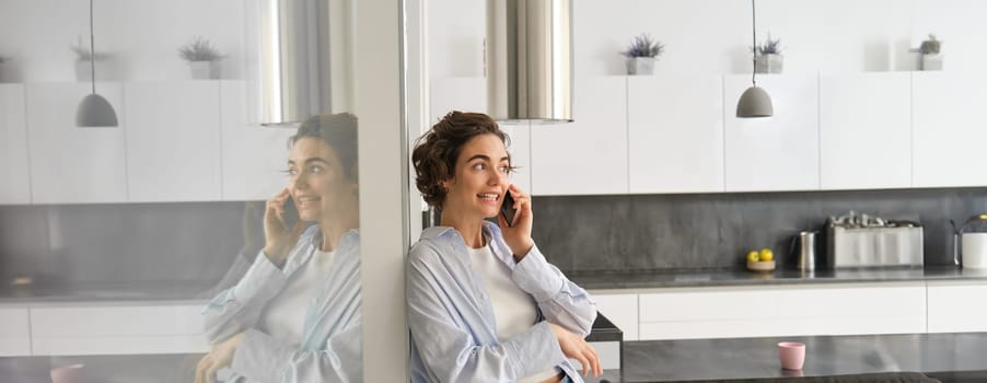 Beautiful woman talking on mobile phone, answers phone call and smiles, stands at home, leans to wall and chats.