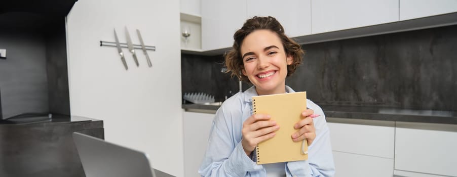 Smiling brunette womnan, holding her diary, writing down in planner, working from home on laptop, studying online and taking notes in notebook.