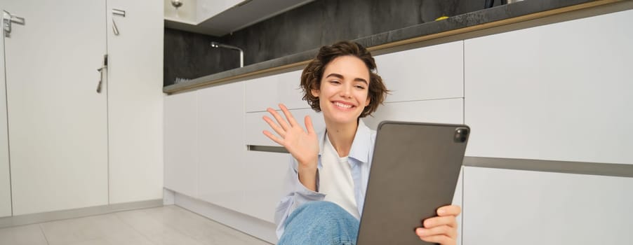 Portrait of happy young woman connects to video chat, waves at digital tablet and smiling, sits at home.