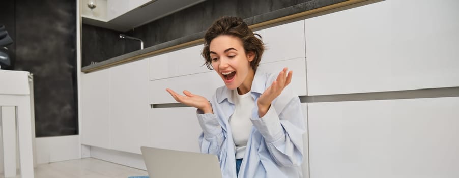 Portrait of young freelance girl connects to online meeting, video chats on laptop, sits on kitchen floor and enjoys her conversation.